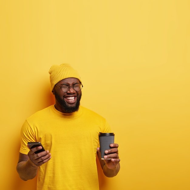 shot happy pleased dark skinned guy closes eyes from pleasure wears yellow stylish hat t shirt messages social networks holds modern cellular coffee go giggles something funny 273609 26213